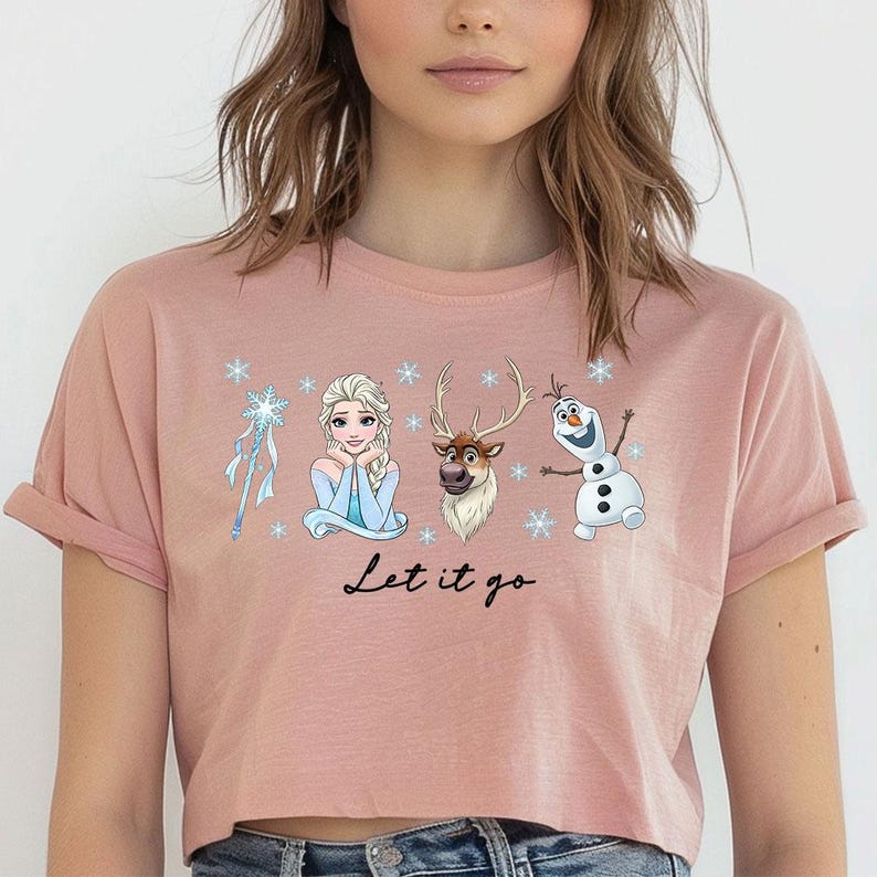May include: A cropped, dusty rose t-shirt featuring a graphic of characters from the movie Frozen, including Elsa, Olaf, and Sven, with the phrase "Let it go" in a script font. The design includes snowflakes and a snowflake wand.