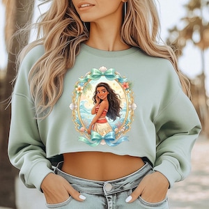 Peut inclure: Un sweat-shirt court vert clair avec une illustration encadr&eacute;e de personnage de dessin anim&eacute;. L'illustration repr&eacute;sente une femme aux cheveux fonc&eacute;s v&ecirc;tue d'une tenue rouge et blanche, sur fond de sc&egrave;ne oc&eacute;anique tropicale. Le cadre est orn&eacute; de fleurs et de n&oelig;uds.