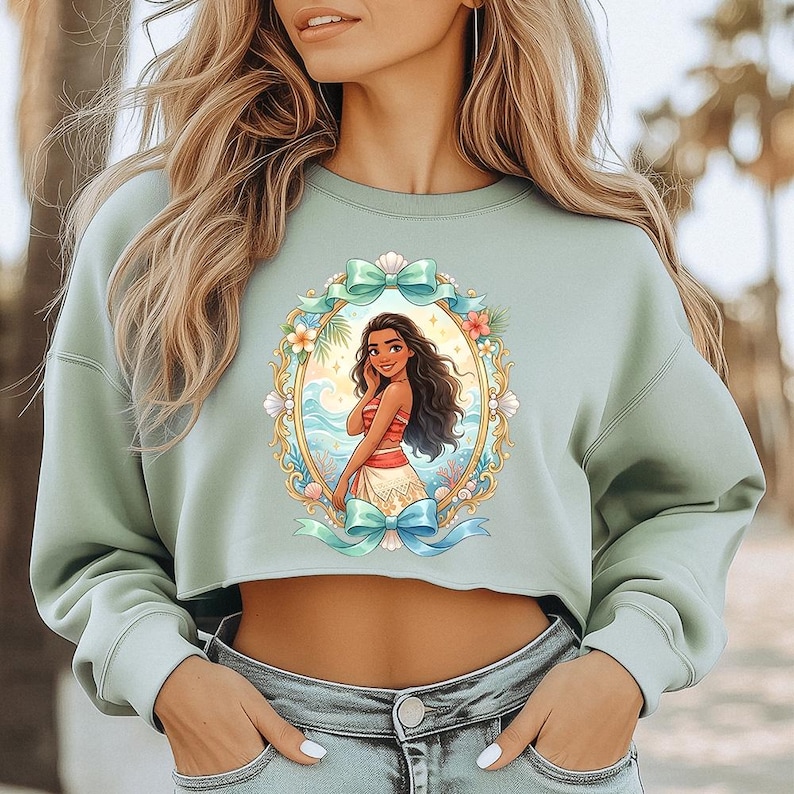 Peut inclure: Un sweat-shirt court vert clair avec une illustration encadr&eacute;e de personnage de dessin anim&eacute;. L'illustration repr&eacute;sente une femme aux cheveux fonc&eacute;s v&ecirc;tue d'une tenue rouge et blanche, sur fond de sc&egrave;ne oc&eacute;anique tropicale. Le cadre est orn&eacute; de fleurs et de n&oelig;uds.