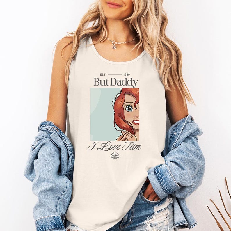 May include: Cream-colored tank top with the text "But Daddy" above an illustration of a cartoon character with red hair and the words "I Love Him". The tank top has a scoop neck and is sleeveless.