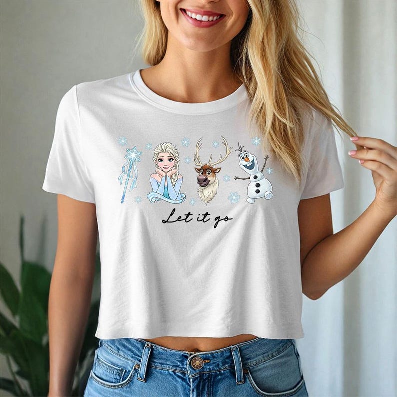May include: White crop top featuring a graphic of Elsa, Sven, Olaf, and a snowflake wand. The text "Let it go" is written in a cursive font below the characters. The shirt is made of a soft material.
