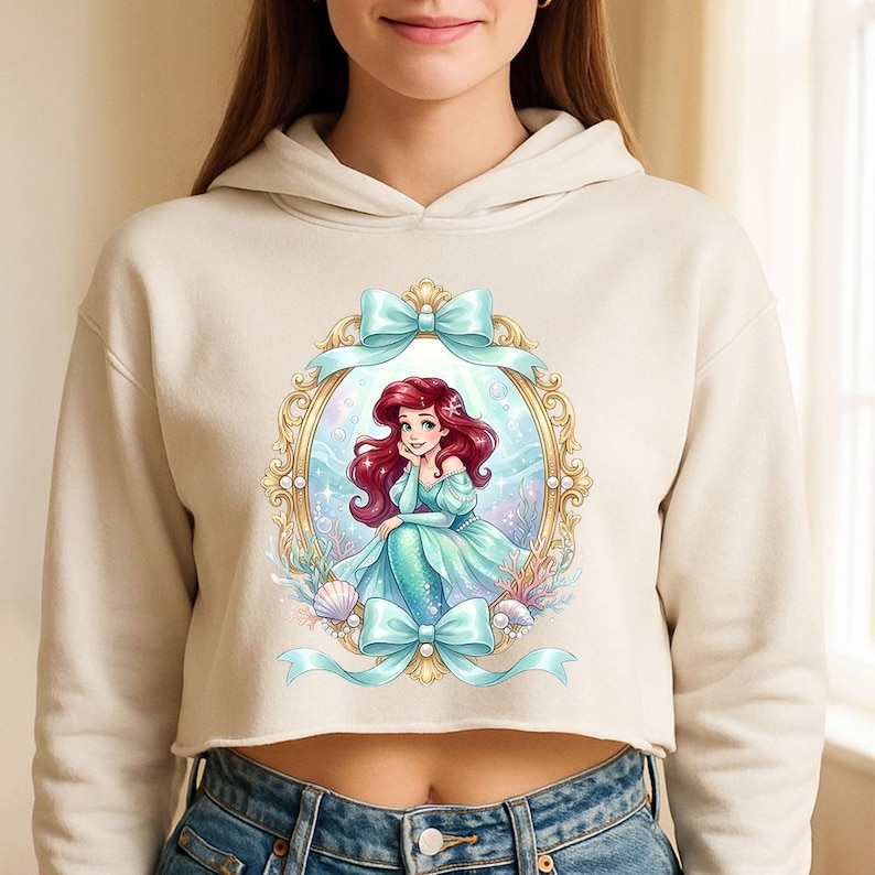 Op de afbeelding: Een cr&egrave;mekleurige cropped hoodie met een afbeelding van Ariel uit De Kleine Zeemeermin. De illustratie toont Ariel in een turquoise jurk en zeemeerminstaart, omlijst door een gouden ovaal en lichtblauwe strikken. Het ontwerp bevat schelpen en waterplanten.