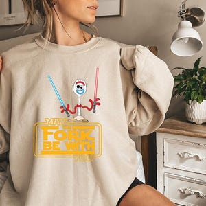 May include: A beige crewneck sweatshirt featuring a graphic of a white spork character with red arms holding a blue and a red lightsaber. Below the character, the yellow text reads "MAY THE FORK BE WITH YOU" inside a rectangular outline.