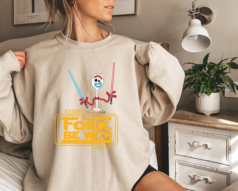May include: A beige crewneck sweatshirt featuring a graphic of a white spork character with red arms holding a blue and a red lightsaber. Below the character, the yellow text reads "MAY THE FORK BE WITH YOU" inside a rectangular outline.
