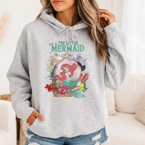 May include: A light gray hooded sweatshirt featuring a graphic print of The Little Mermaid characters including Ariel in a shell, King Triton, Ursula, Sebastian, and Flounder. The design includes colorful underwater elements and bubbles.
