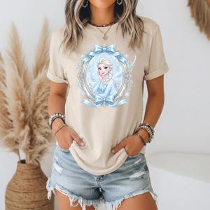 May include: Beige t-shirt with a cartoon illustration of a princess in a blue dress, framed by a blue ribbon and an ornate oval border. The princess has blonde hair and is smiling. The shirt has short sleeves.