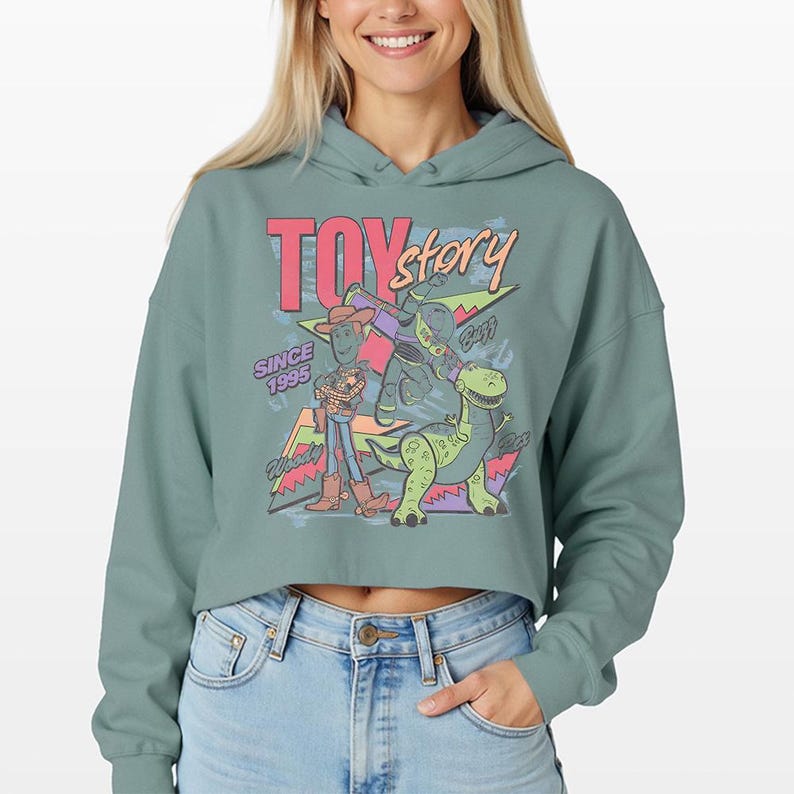 May include: A sage green cropped hoodie featuring a graphic print of Toy Story characters Woody, Buzz Lightyear, and Rex. The design includes the text "TOY story" in pink and orange, "SINCE 1995" in purple, and "Woody", "Buzz", and "Rex" in script.