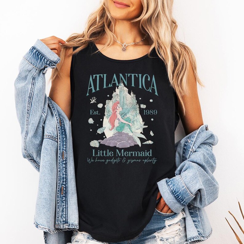Op de afbeelding: Zwart mouwloos shirt met een afbeelding van de Kleine Zeemeermin, de tekst "Atlantica" en het jaar "1989". Het ontwerp bevat een kasteelillustratie en de zin "We have gadgets & gizmos aplenty".