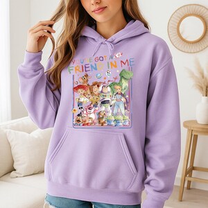 May include: A light purple hoodie featuring a colorful graphic of characters from the movie Toy Story. The graphic includes the text "YOU'VE GOT A FRIEND IN ME". The hoodie has a front pocket and a drawstring hood.