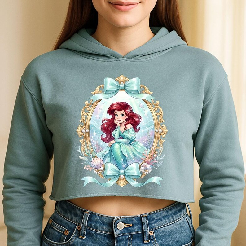 Op de afbeelding: Een lichtblauwe cropped hoodie met een ingelijste illustratie van een zeemeermin met rood haar en een blauwgroene staart. De illustratie is omgeven door blauwgroene strikken, schelpen en koraal. De hoodie heeft een capuchon met trekkoord en lange mouwen.