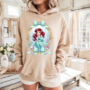 Puede incluir: Sudadera con capucha beige con una colorida ilustraci&oacute;n de una sirena dentro de un marco ovalado dorado con lazos azul claro. La sirena tiene el pelo rojo y lleva un vestido azul claro. La sudadera tiene un bolsillo de canguro y una capucha con cord&oacute;n.