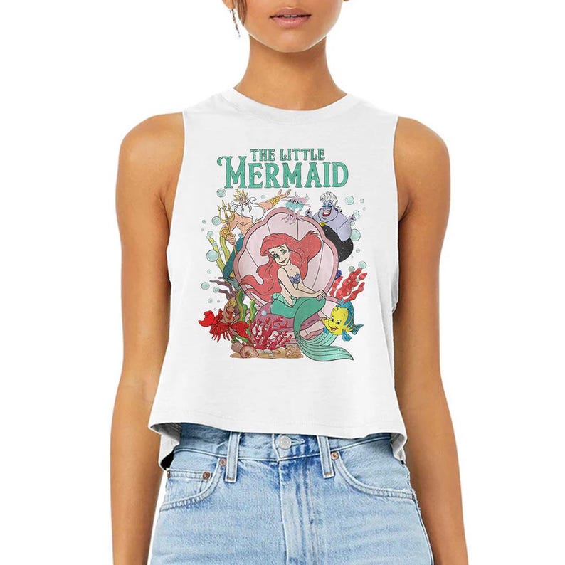 Puede incluir: Un top corto sin mangas de color blanco con un estampado gr&aacute;fico colorido de los personajes de La Sirenita, incluyendo a Ariel, el Rey Trit&oacute;n, &Uacute;rsula, Sebasti&aacute;n y Flounder. El dise&ntilde;o incluye elementos submarinos como coral, burbujas y una gran concha.