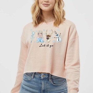 May include: A light pink cropped sweatshirt featuring characters from the movie Frozen. The graphic includes Elsa, Olaf, Sven, and a snowflake wand. The phrase "Let it go" is written in black script below the characters.
