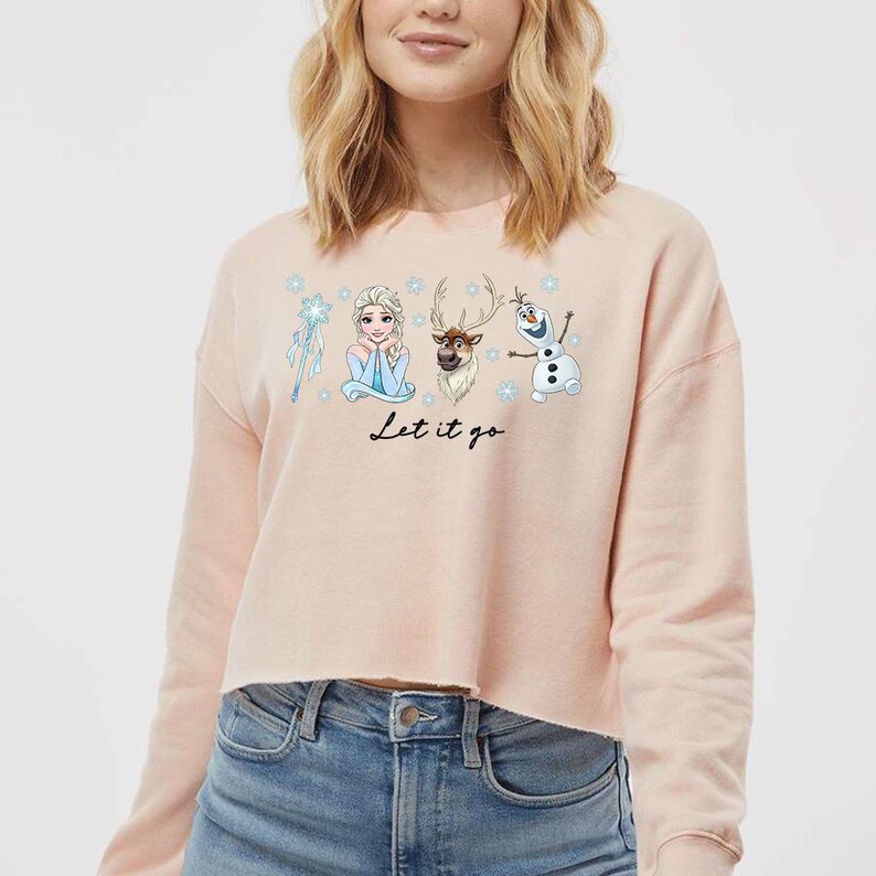 May include: A light pink cropped sweatshirt featuring characters from the movie Frozen. The graphic includes Elsa, Olaf, Sven, and a snowflake wand. The phrase "Let it go" is written in black script below the characters.