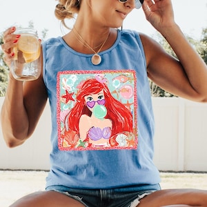 May include: Light blue tank top featuring a colorful graphic of a cartoon mermaid with red hair, heart-shaped sunglasses, and a bubble. The graphic is framed with a pink border and includes seashells and starfish.