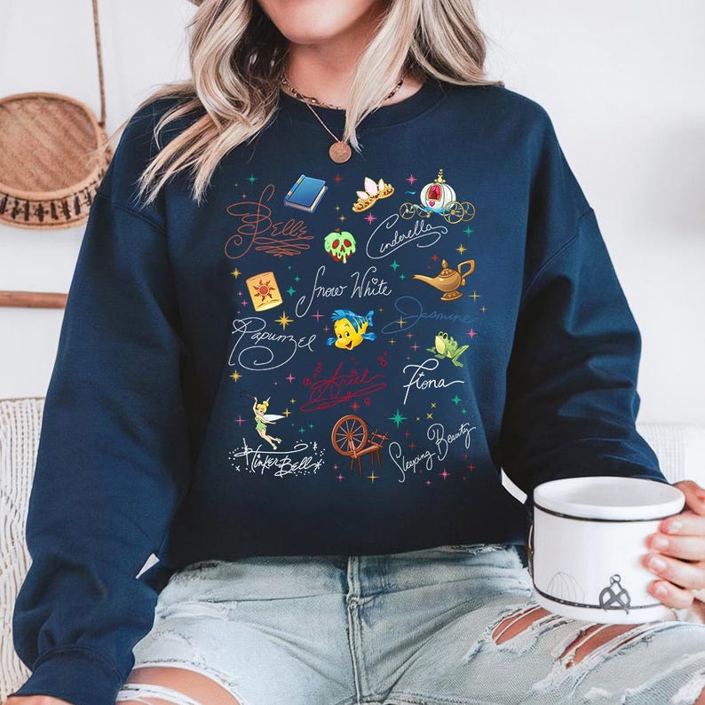 May include: A navy blue crewneck sweatshirt featuring a colorful graphic print of various fairy tale icons and character names including Belle, Cinderella, Snow White, Jasmine, Rapunzel, Fiona, Tinker Bell, and Sleeping Beauty with small stars.