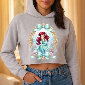 Op de afbeelding: Een lichtgrijze cropped hoodie met een ingelijste illustratie van een zeemeermin met rood haar en een turquoise jurk. Het gouden frame heeft turquoise strikken. De zeemeermin is omringd door schelpen en waterplanten.