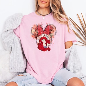 Puede incluir: Camiseta rosa claro con un gr&aacute;fico de Ariel de La Sirenita. El dise&ntilde;o incluye una cabeza estilo Minnie Mouse con un lazo rojo de lunares y la cara de Ariel. La camiseta es de color liso y tiene mangas cortas.