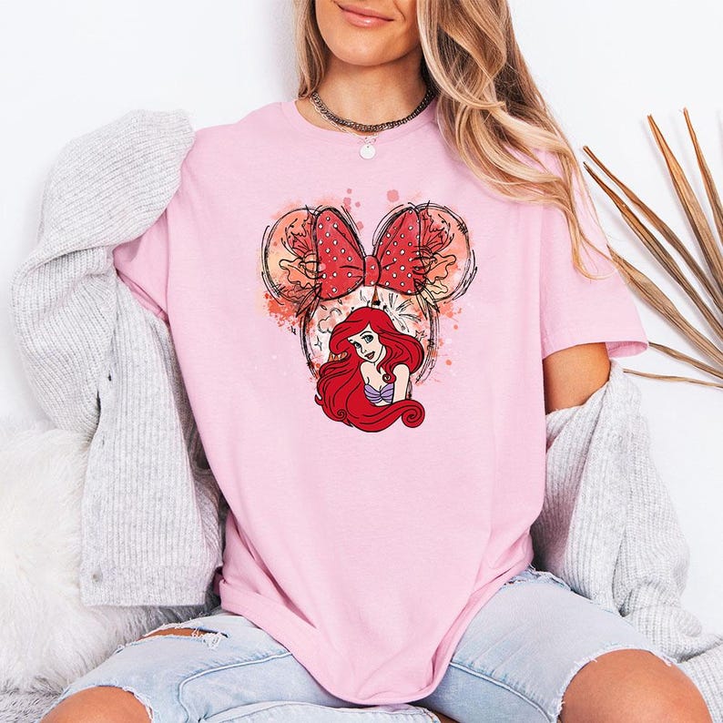 Puede incluir: Camiseta rosa claro con un gr&aacute;fico de Ariel de La Sirenita. El dise&ntilde;o incluye una cabeza estilo Minnie Mouse con un lazo rojo de lunares y la cara de Ariel. La camiseta es de color liso y tiene mangas cortas.