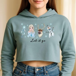May include: A cropped, teal hoodie with illustrations of characters from the film Frozen, including Elsa, Olaf, and Sven, with snowflakes and the text "Let it go". The hoodie has long sleeves and a drawstring hood.