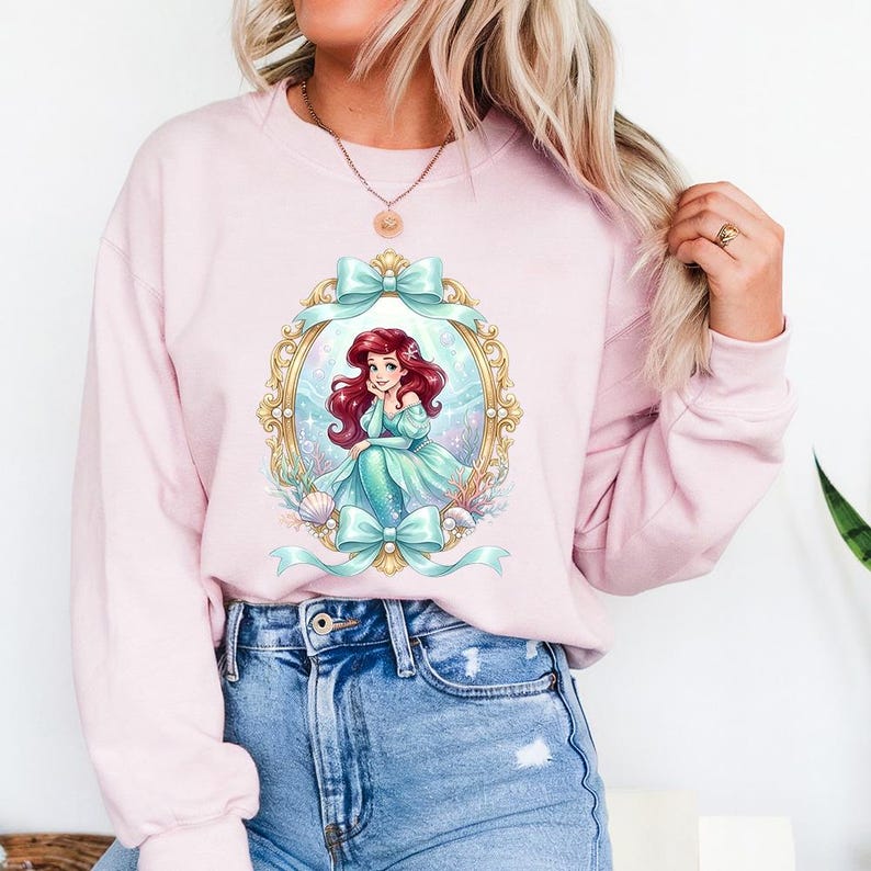 May include: A light pink sweatshirt featuring a framed illustration of a mermaid with red hair and a teal dress. The frame is gold with teal bows. The sweatshirt is a long-sleeved crew neck.