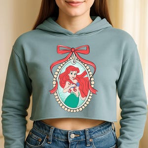 Puede incluir: Una sudadera corta de color verde azulado claro con una ilustraci&oacute;n enmarcada de Ariel de La Sirenita. La ilustraci&oacute;n est&aacute; rodeada por un borde de perlas y un lazo rojo. Ariel tiene el pelo rojo, un top azul y una cola verde.