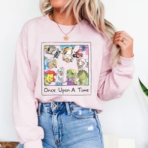 May include: Light pink sweatshirt featuring a framed graphic with cartoon characters and the text "Once Upon A Time". The graphic includes various animated characters. The sweatshirt is a solid light pink color.