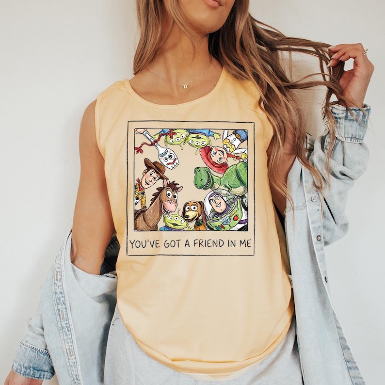 May include: Pale yellow sleeveless top with a framed graphic of Toy Story characters, including Woody, Buzz Lightyear, and the Little Green Men. The phrase "YOU'VE GOT A FRIEND IN ME" is printed below the image. The top is made of a soft material.
