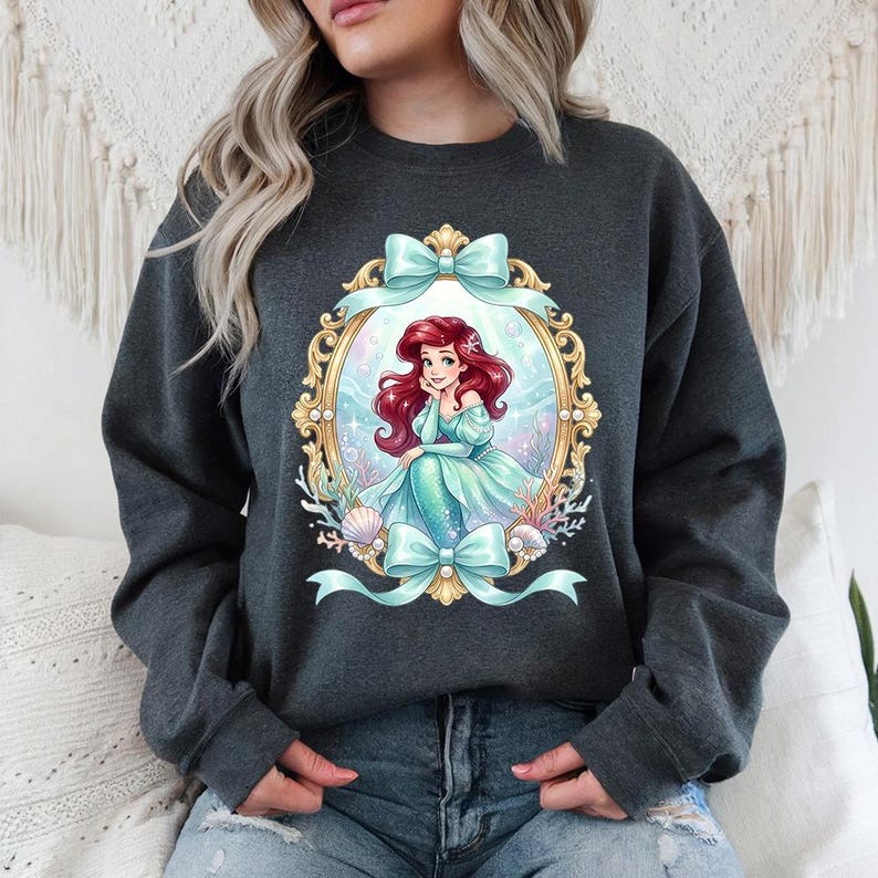 May include: Dark gray sweatshirt featuring a framed illustration of a mermaid with red hair and a teal dress. The frame is gold with teal bows. The mermaid is surrounded by seashells and coral.