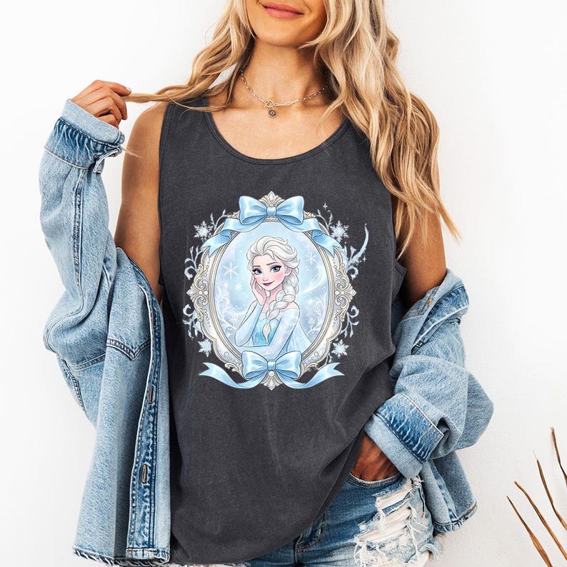 May include: Dark gray tank top featuring a framed illustration of a cartoon character with white hair and a blue dress. The frame is decorated with blue bows and snowflakes. The tank top is sleeveless.