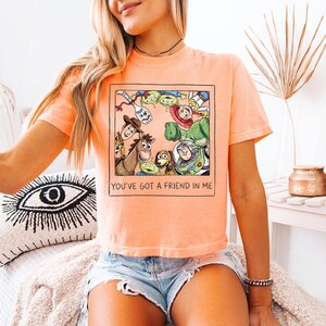 May include: Orange t-shirt with a framed graphic of Toy Story characters, including Woody and Buzz Lightyear. The graphic is surrounded by a black border. The text "YOU'VE GOT A FRIEND IN ME" is printed below the graphic.