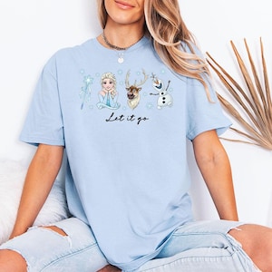 May include: Pale blue t-shirt with a graphic of Elsa, a reindeer, and Olaf from the film Frozen. The words "Let it go" are written in black script below the characters. The shirt is made of a soft, comfortable material.
