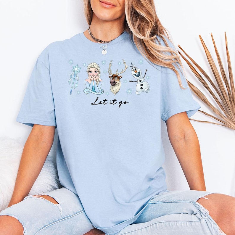May include: Pale blue t-shirt with a graphic of Elsa, a reindeer, and Olaf from the film Frozen. The words "Let it go" are written in black script below the characters. The shirt is made of a soft, comfortable material.