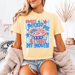 Könnte beinhalten: Ein hellgelbes Kurzarm-T-Shirt mit einem Grafikdruck einer Torte, die mit einem amerikanischen Flaggenmuster verziert ist. Der Text lautet SWEET POTATO PIE & I SHUT MY MOUTH in blauen und roten Buchstaben, umgeben von blauen und roten Sternen.