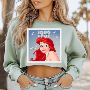 Pode incluir: Camisola curta verde claro com um desenho da Ariel de A Pequena Sereia. O desenho inclui o texto "1989 Ariel's Version" e uma ilustra&ccedil;&atilde;o da Ariel com gaivotas num fundo azul.