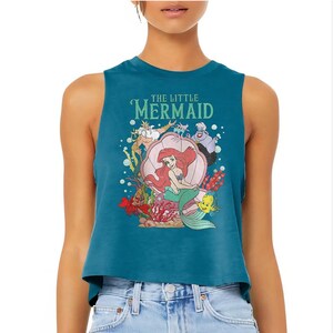 Puede incluir: Un top corto sin mangas de color azul cerceta con un estampado gr&aacute;fico de los personajes de La Sirenita, incluyendo a Ariel sentada en una concha, el Rey Trit&oacute;n, &Uacute;rsula, Sebasti&aacute;n y Flounder, rodeados de burbujas y corales.