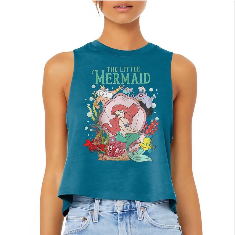 Puede incluir: Un top corto sin mangas de color azul cerceta con un estampado gr&aacute;fico de los personajes de La Sirenita, incluyendo a Ariel sentada en una concha, el Rey Trit&oacute;n, &Uacute;rsula, Sebasti&aacute;n y Flounder, rodeados de burbujas y corales.