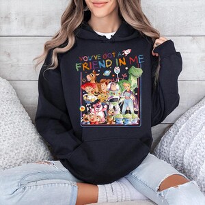 May include: A navy blue hooded sweatshirt featuring a colorful graphic of characters from a popular animated movie. The text "YOU'VE GOT A FRIEND IN ME" is printed above the graphic. The sweatshirt is worn by a person.