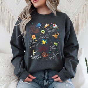May include: A dark gray crewneck sweatshirt featuring a graphic print of various fairy tale icons and character names in cursive script, including Belle, Cinderella, Snow White, Jasmine, Rapunzel, Fiona, Tinker Bell, and Sleeping Beauty.