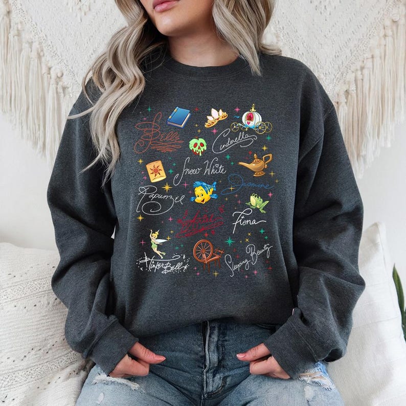 May include: A dark gray crewneck sweatshirt featuring a graphic print of various fairy tale icons and character names in cursive script, including Belle, Cinderella, Snow White, Jasmine, Rapunzel, Fiona, Tinker Bell, and Sleeping Beauty.