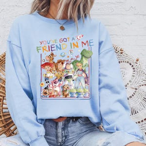 May include: Light blue sweatshirt featuring a colorful graphic of characters from the animated film "Toy Story." The graphic includes Woody, Buzz Lightyear, and other characters, with the text "You've Got a Friend in Me."