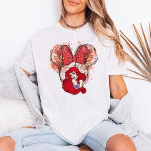 Puede incluir: Camiseta blanca con un gr&aacute;fico de Ariel de La Sirenita. El dise&ntilde;o incluye un lazo rojo con lunares, orejas de Mickey Mouse y detalles de acuarela. Ariel tiene el pelo largo rojo y un top de concha p&uacute;rpura.