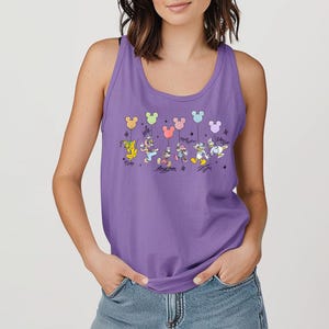 May include: A lilac tank top with a vibrant graphic of Disney characters, including Pluto, Mickey, Minnie, Donald, Daisy, and Goofy, holding Mickey Mouse-shaped balloons. Each character's signature is below. The top is sleeveless with a scoop neckline.