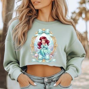 Puede incluir: Una sudadera corta verde salvia con una ilustraci&oacute;n enmarcada de una sirena con cabello rojo y un vestido turquesa. La ilustraci&oacute;n est&aacute; rodeada de lazos y conchas marinas turquesas. La sudadera tiene mangas largas y un corte holgado.