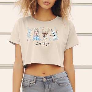 May include: A beige crop top featuring a graphic of characters from the movie Frozen, including Elsa, Olaf, and Sven, with the text "Let it go" in a script font. The shirt has short sleeves and a crew neck.