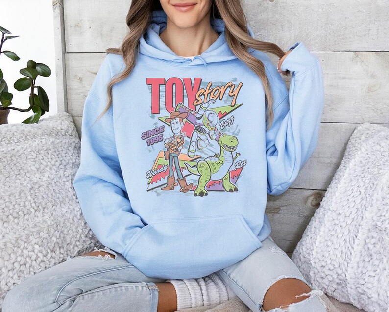 May include: A light blue hooded sweatshirt featuring a graphic print of Toy Story characters Woody, Buzz Lightyear, and Rex. The design includes the text Toy Story, Since 1995, Woody, Buzz, and Rex in a colorful, retro-inspired font and layout.