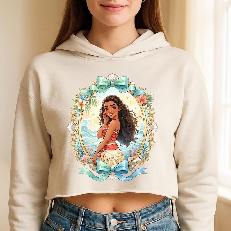 K&ouml;nnte beinhalten: Cremefarbener, verk&uuml;rzter Kapuzenpullover mit einer farbenfrohen Illustration einer Zeichentrickfigur in einem verzierten ovalen Rahmen. Der Rahmen ist mit Blumen, Muscheln und t&uuml;rkisfarbenen Schleifen verziert. Die Figur hat lange dunkle Haare und tr&auml;gt ein rot-wei&szlig;es Outfit.