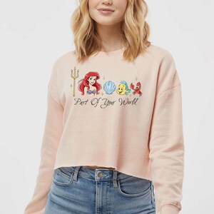 May include: A pale pink cropped sweatshirt with a graphic of Ariel, a seashell, Flounder, and Sebastian from The Little Mermaid. The text "Part Of Your World" is below the graphic. The sweatshirt is worn with blue jeans.