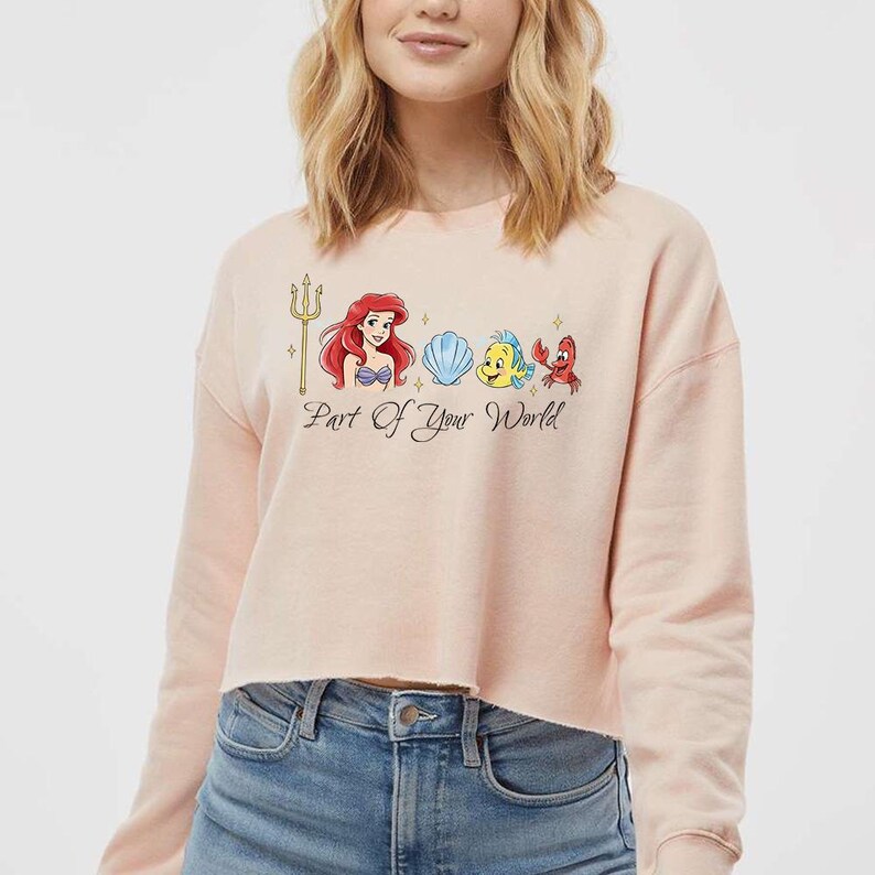 May include: A pale pink cropped sweatshirt with a graphic of Ariel, a seashell, Flounder, and Sebastian from The Little Mermaid. The text "Part Of Your World" is below the graphic. The sweatshirt is worn with blue jeans.
