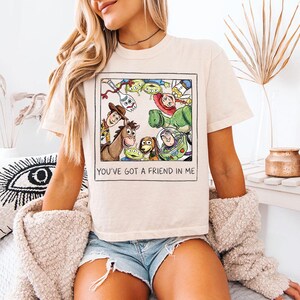 May include: A cream-coloured t-shirt with a rectangular graphic featuring cartoon characters from the film Toy Story. The graphic includes Woody, Buzz Lightyear, and other characters. The text "YOU'VE GOT A FRIEND IN ME" is below the graphic.